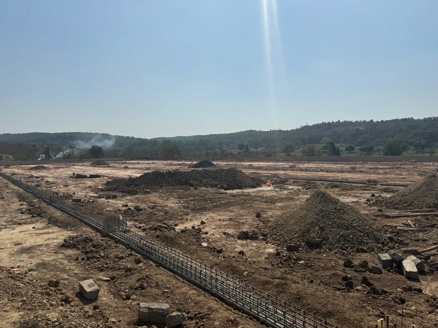 Foundations and reinforcement laid out across the cleared farm site at Kulavalli, with wooded hills behind